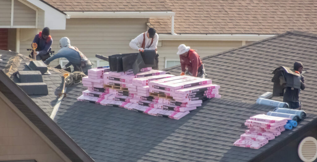 Roofers installing shingles on a residential roof, with stacks of roofing materials visible.