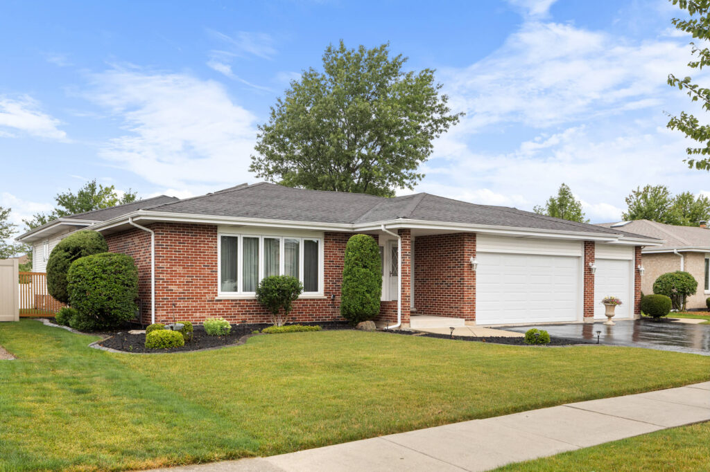 suburban home with a shingle roof, brick siding, a three car garage, neat landscaping, and a wet blacktop driveway.
